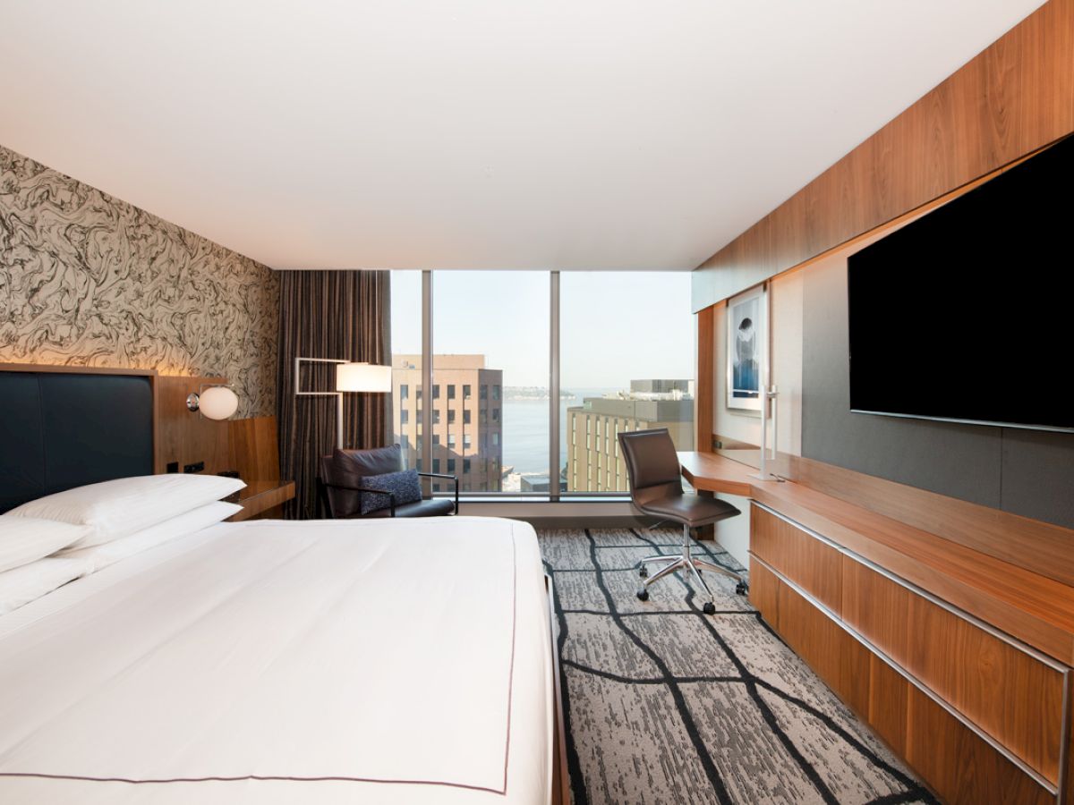 A modern hotel room with a large bed, wall-mounted TV, work desk and chair, and a window view of city buildings and water is shown.