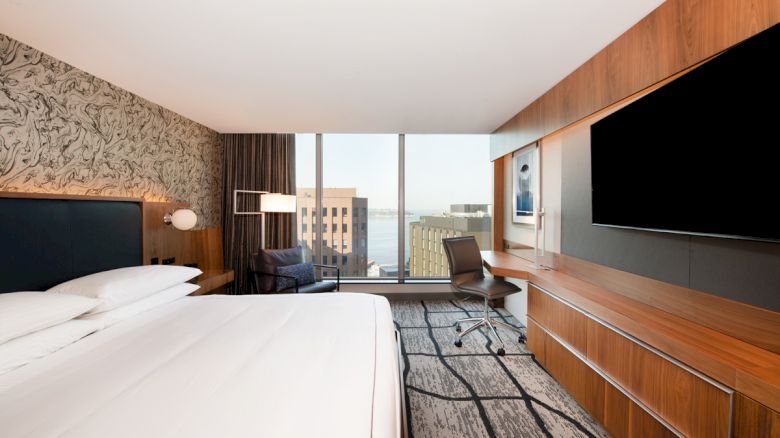A modern hotel room with a large bed, wall-mounted TV, work desk and chair, and a window view of city buildings and water is shown.