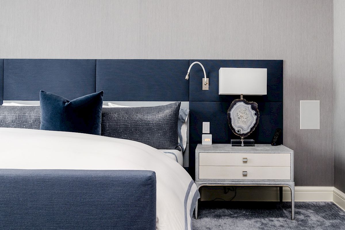 A modern bedroom with a blue upholstered headboard, pillows, white bedding, and a nightstand with a lamp on a soft carpeted floor.