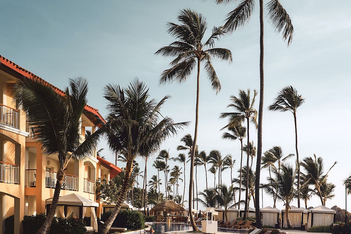 A tropical resort poolside scene with lounge chairs, palm trees, and a building under a clear blue sky.
