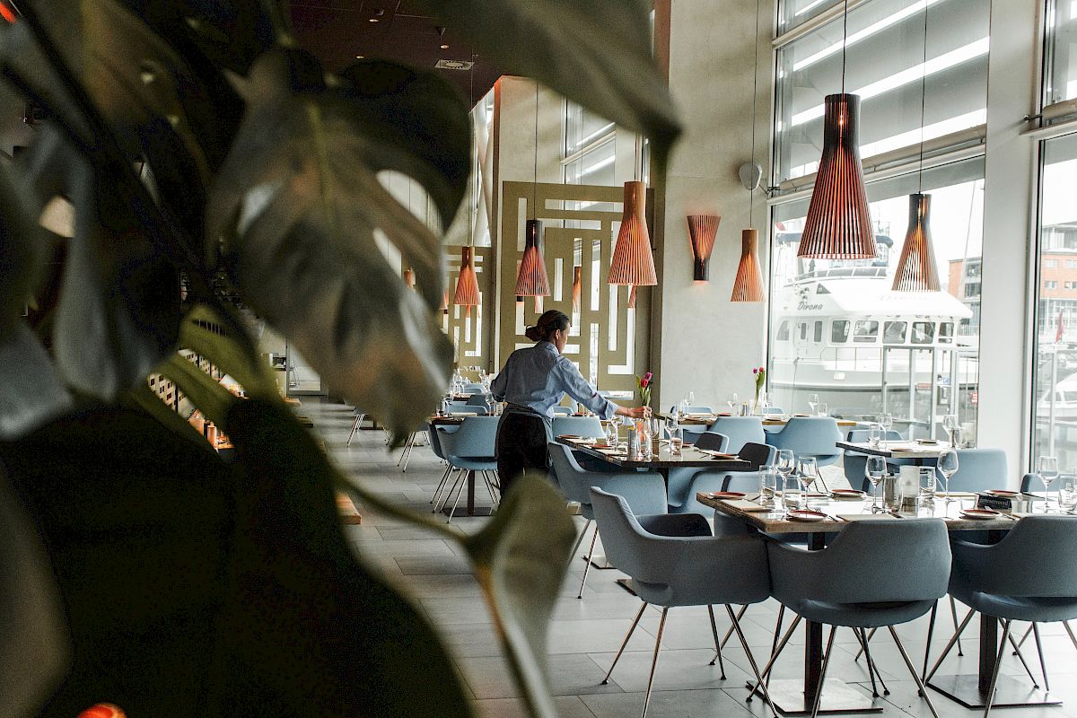The image shows a modern restaurant interior with a person setting tables, featuring stylish lighting and a leafy plant in the foreground.