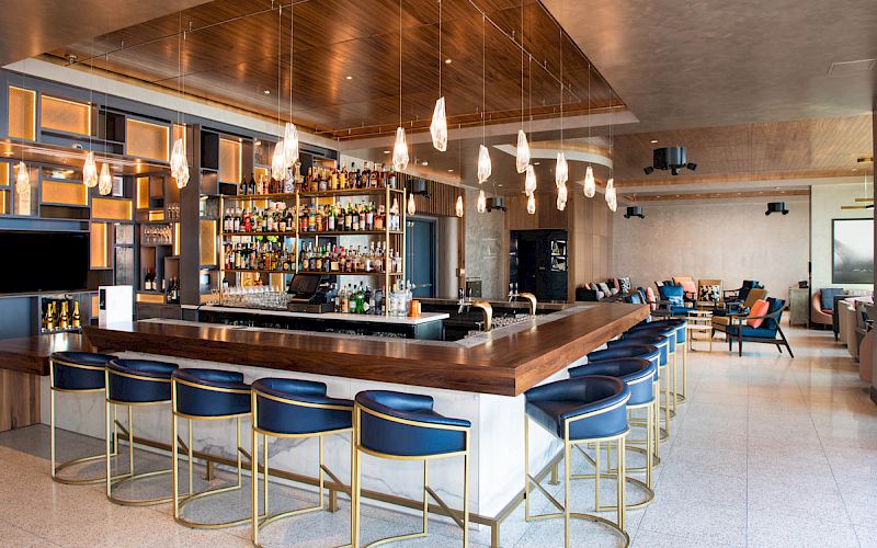 A stylish bar with a wooden countertop, blue chairs, a variety of bottles on shelves, pendant lights, and a cozy seating area in the background.